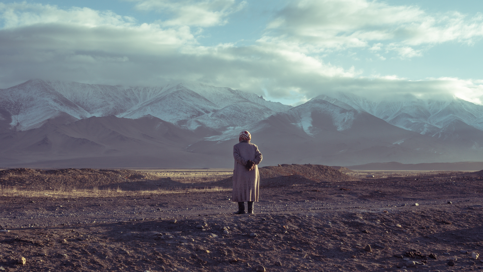 Osamělá starší žena stojí na prašné pláni zády ke kameře a kouká na vysoké zasněžené hory.