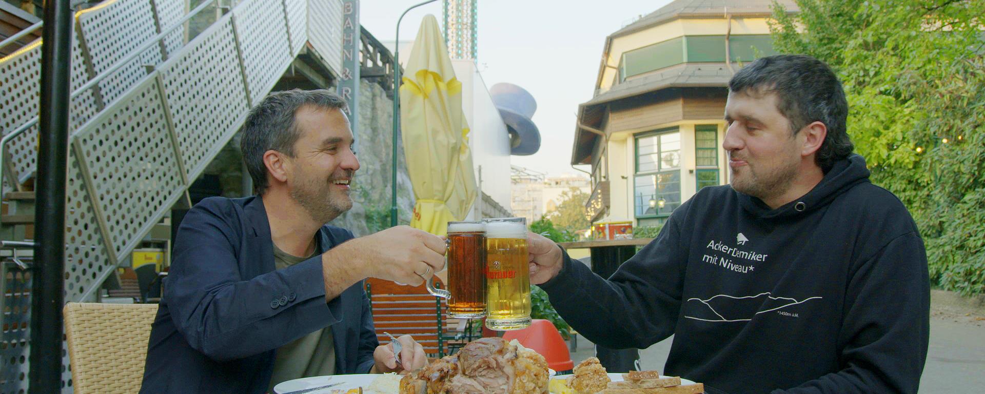 Dva muži sedí u stolu a ťukají si velkým pivem. Na stole jsou hranolky a uzené koleno.