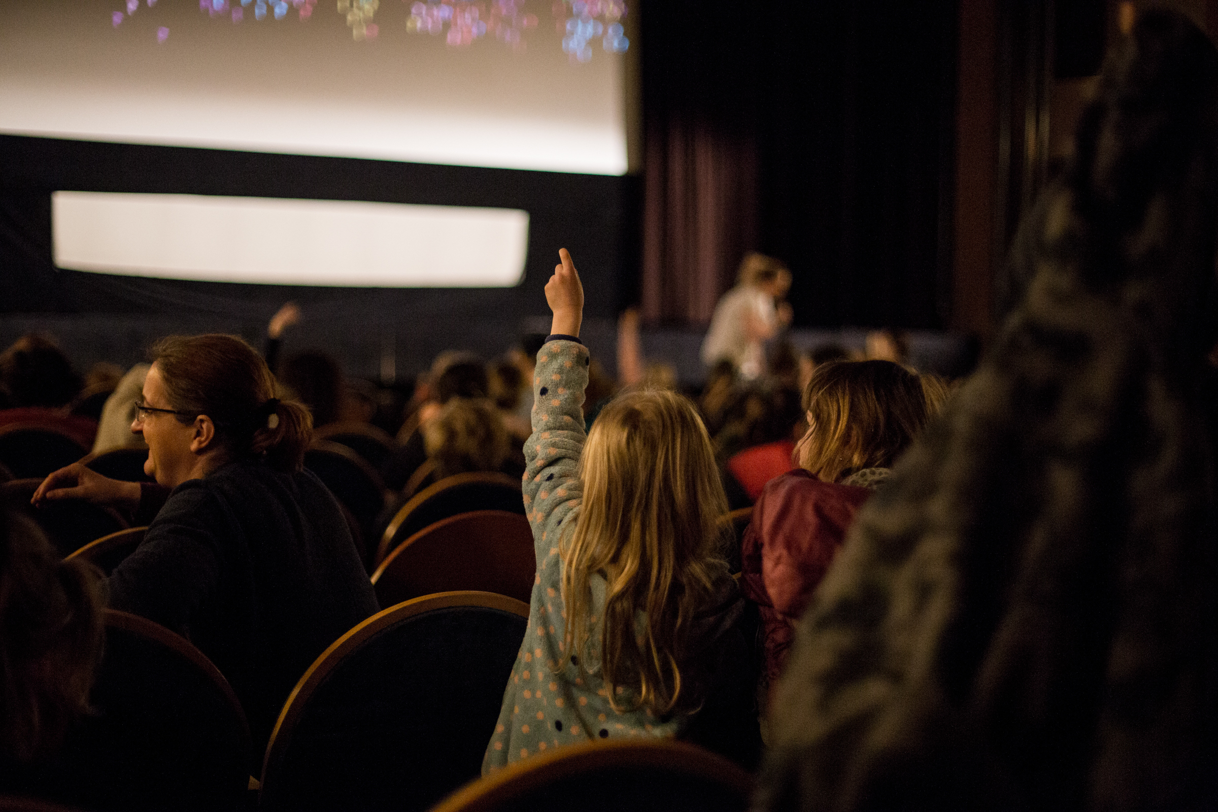 Děti v kině zvedají ruce a povídají si o filmu. V sále je tlumené osvětlení.