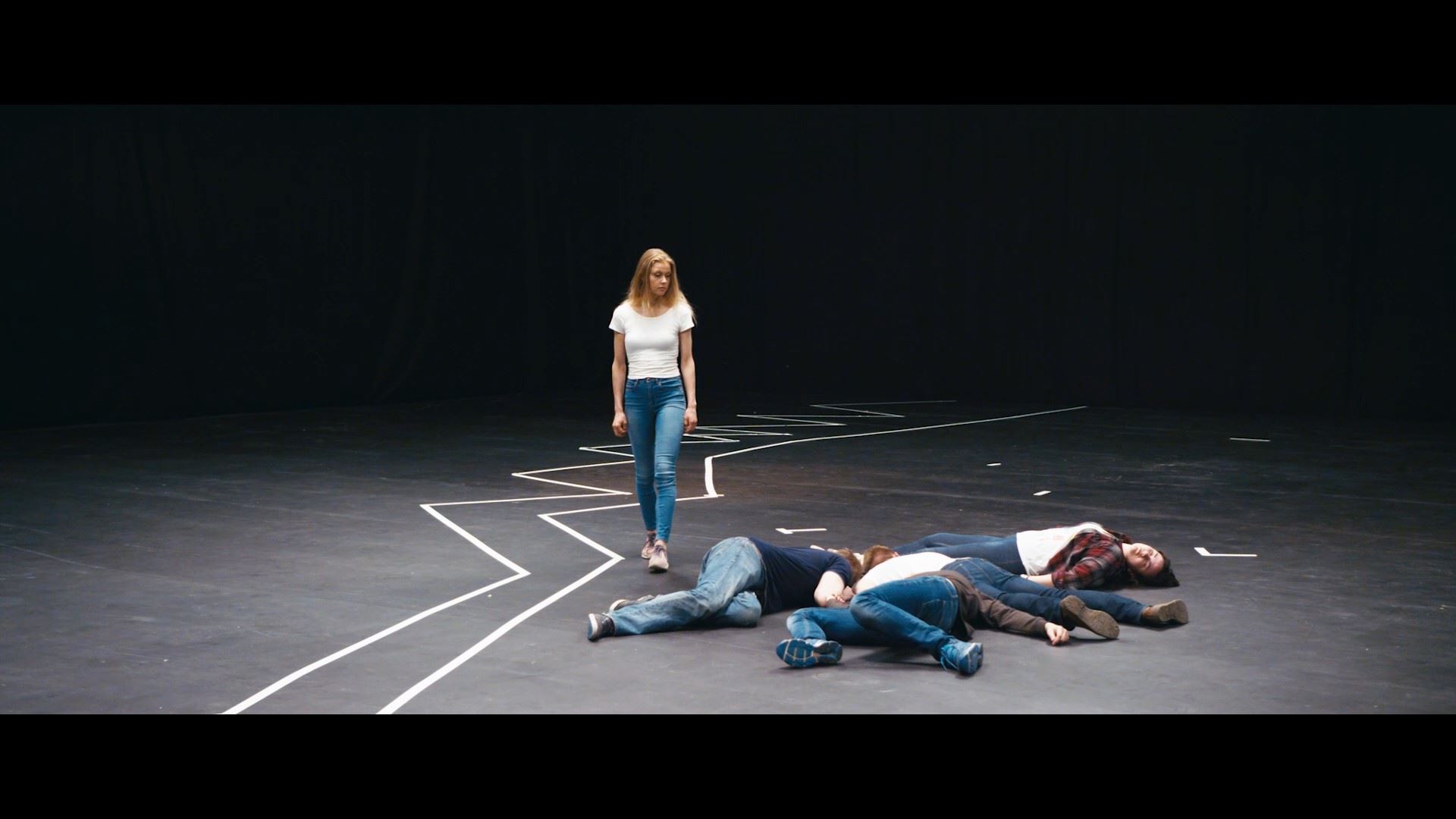 Na scéně bez pozadí stojí osoba a vedle ní leží na zemi čtyři osoby. Na podlaze jsou lepící páskou vyznačené různé linie.