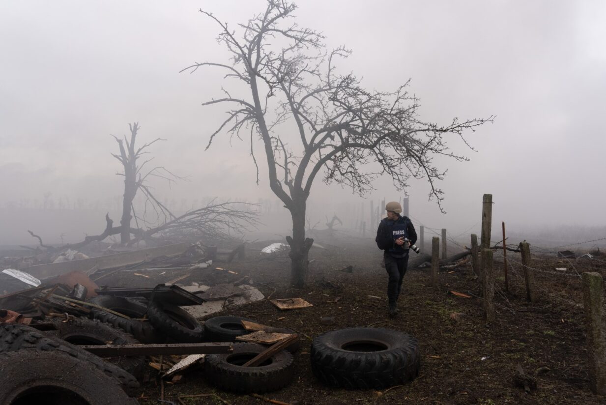 Potemnělá krajina v kouři, ohořelé stromy, zátarasa z pneumatik a ostnatého drátu. Krajinou prochází novinář v helmě s fotoaparátem v ruce.