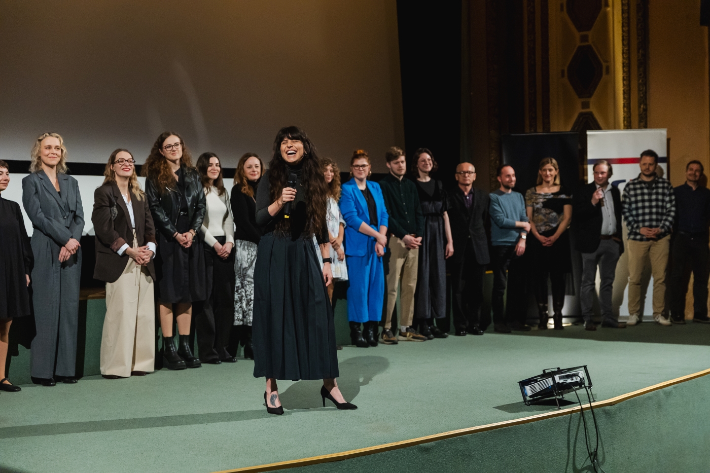 Skupina lidí stojí na pódiu, jedna žena uprostřed mluví do mikrofonu.