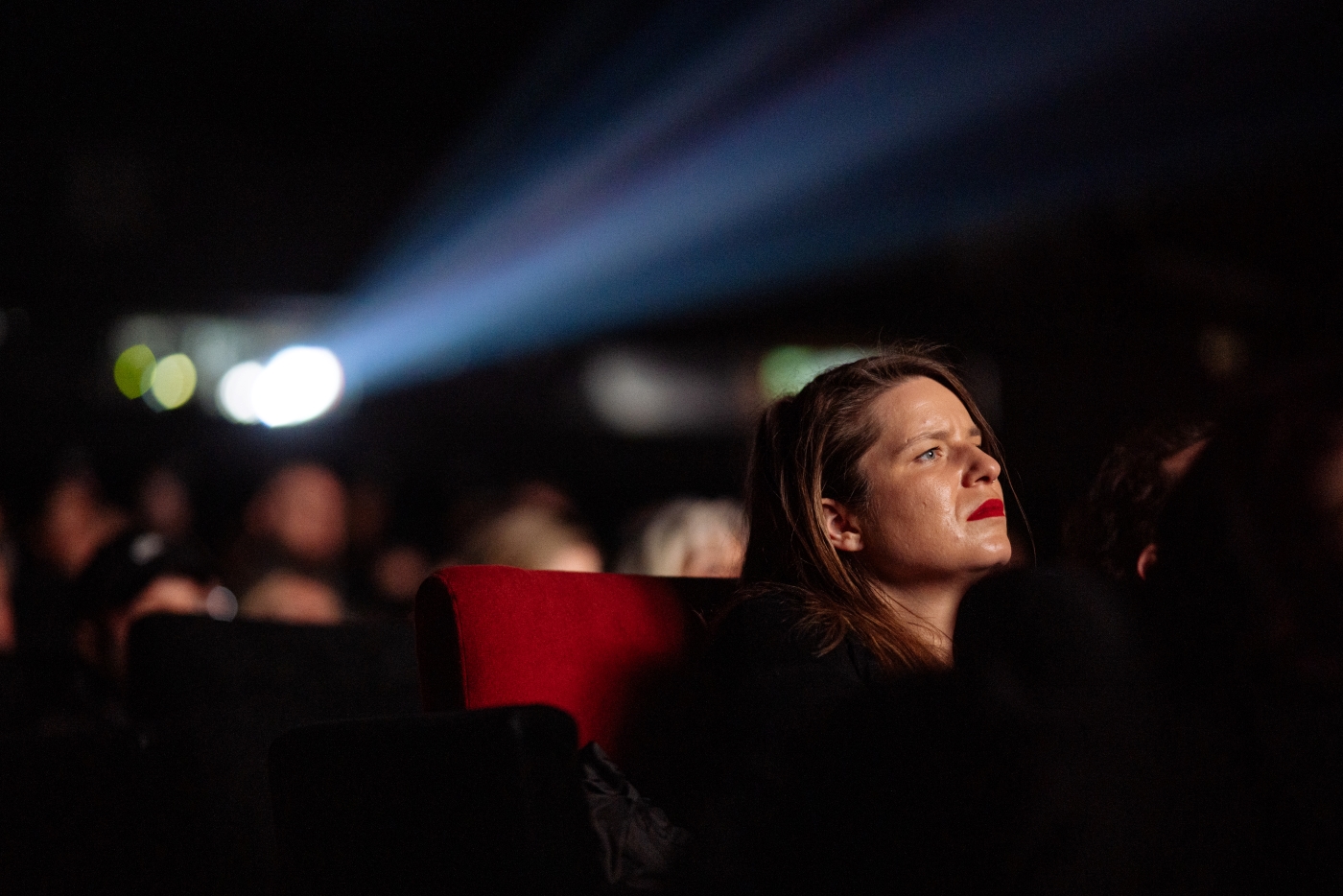 Žena sedí v kině a sleduje film, za ní je vidět světelný paprsek z projektoru.