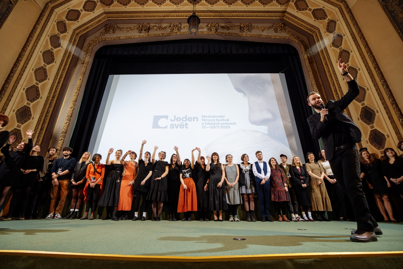 Skupina lidí stojí na pódiu před plátnem s logem festivalu Jeden svět, jeden muž mluví do mikrofonu.