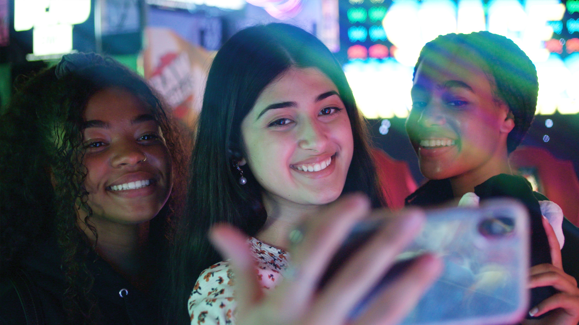 Tři mladé ženy si fotí tzv selfie. Usmívají se, za nimi svítí neony a billboardy města. / Three young women are taking selfies. They are smiling, with the neon and billboards of the city lit up behind them.